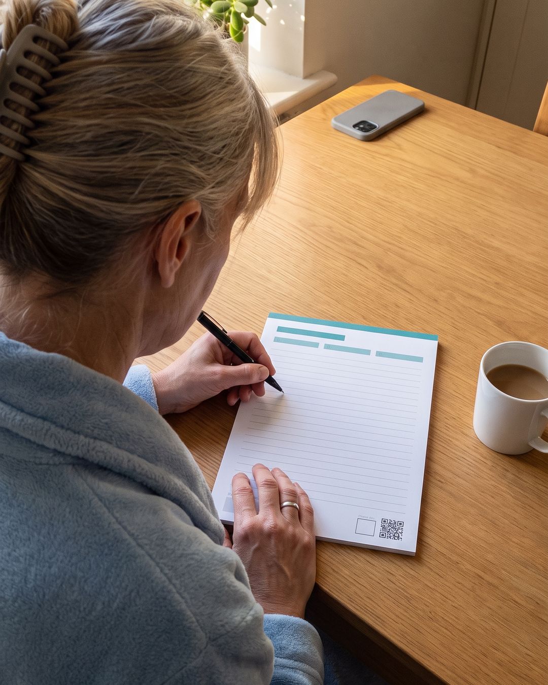 A printed Pen Before Phone page on a kitchen table, pen at the start of the first line, phone face down beside a mug